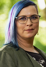 Erin has shoulder-length hair dyed a playful blue and purple and wears glasses, earrings, and a green shirt.