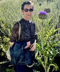 Mimi has short black hair and wears red sunglasses, green earrings, and a patterned black shirt. She stands next to a tall, spiky plant with a purple flower.