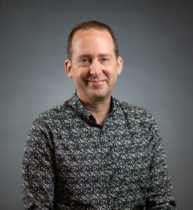 Ken has brown hair, wears a patterned button-up shirt, and is smiling.