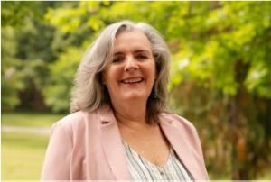 Saskia smiles against a background of vibrant green trees. She has shoulder-length grey-brown hair and wears a striped shirt under a light pink blazer.