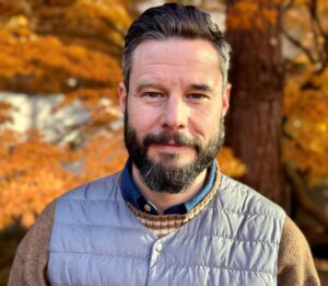 Will has short dark hair and a beard and wears a grey vest over an orange and brown sweater that matches the autumn leaves in the background.