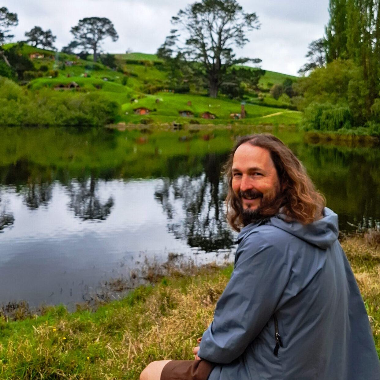 Lee smiles over his shoulder in front of a lake, green hills, and trees at Hobbiton. He has wavy shoulder-length hair and a beard and wears a blue jacket.